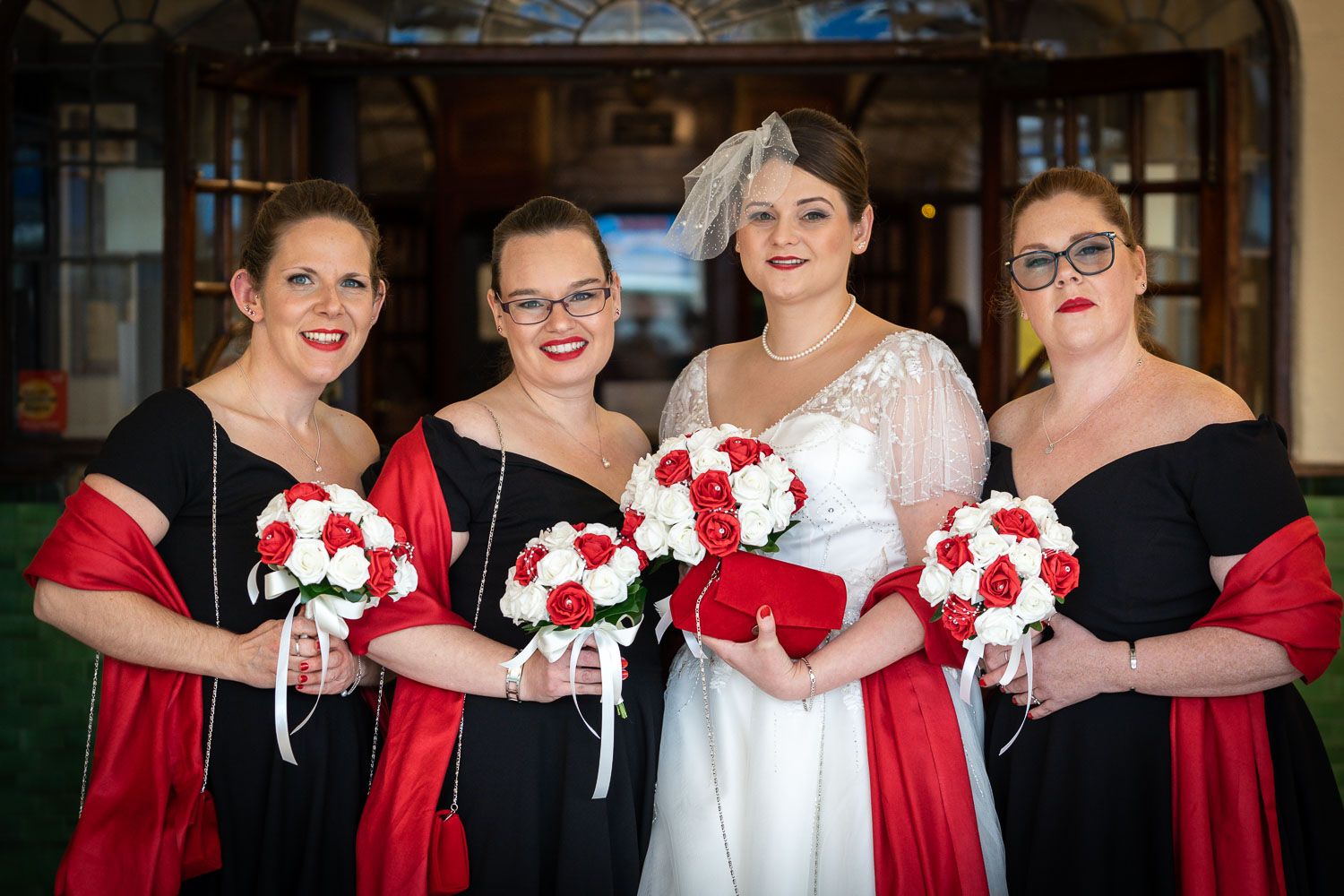 Bridal party poses together wearing elegant black dresses with red accessories and holding red and white bouquets.