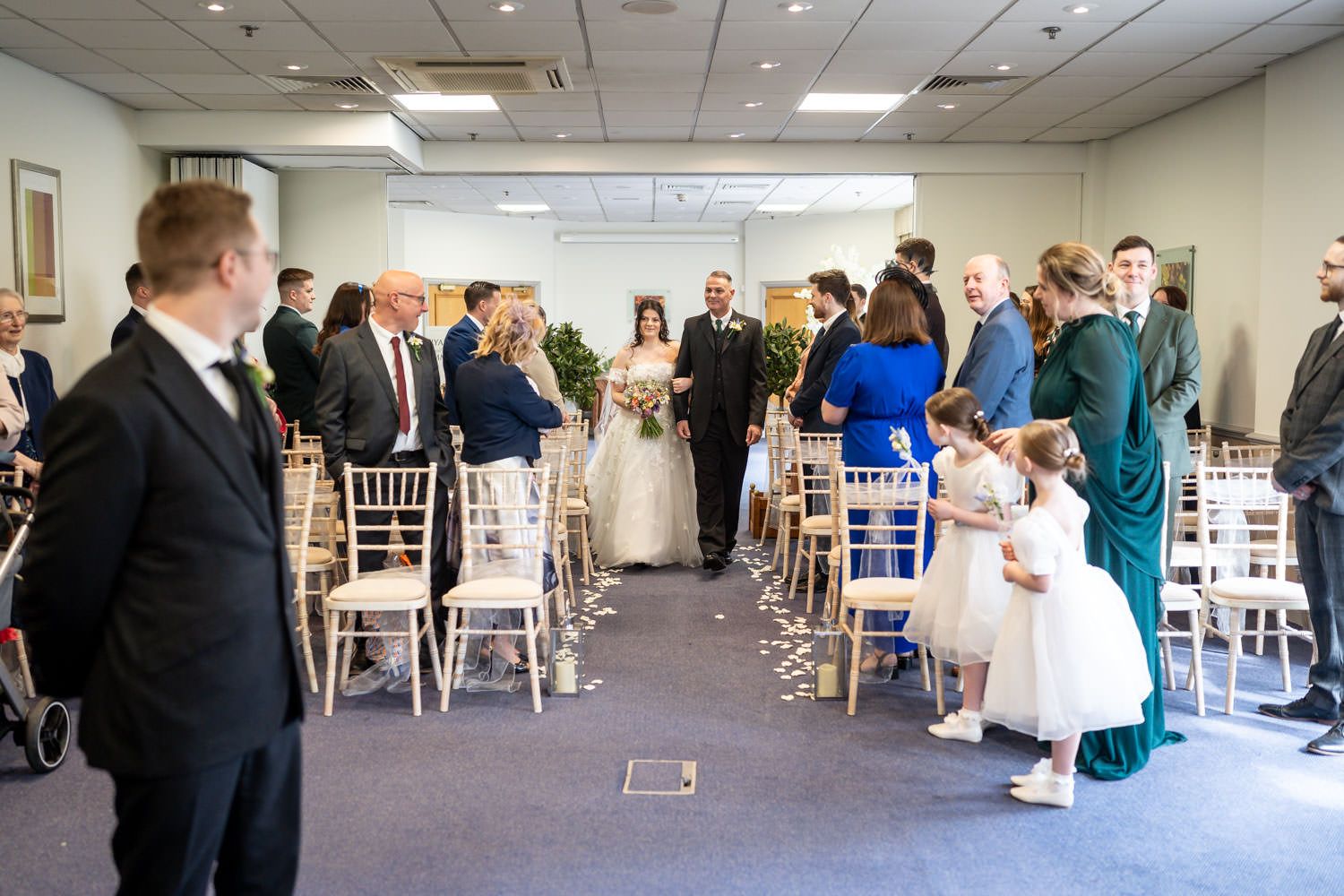 Wedding guests gather in a reception room with white chiavari chairs and floral petals scattered down the aisle.
