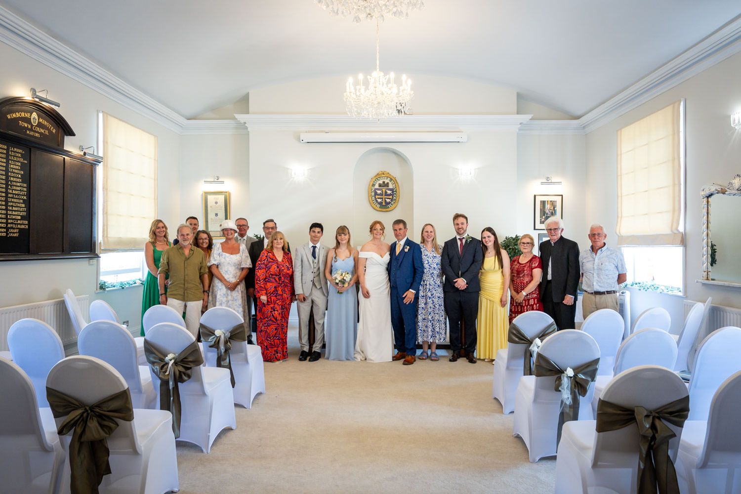 Wedding party and family group photo inside an elegant white ceremony room with chandelier.