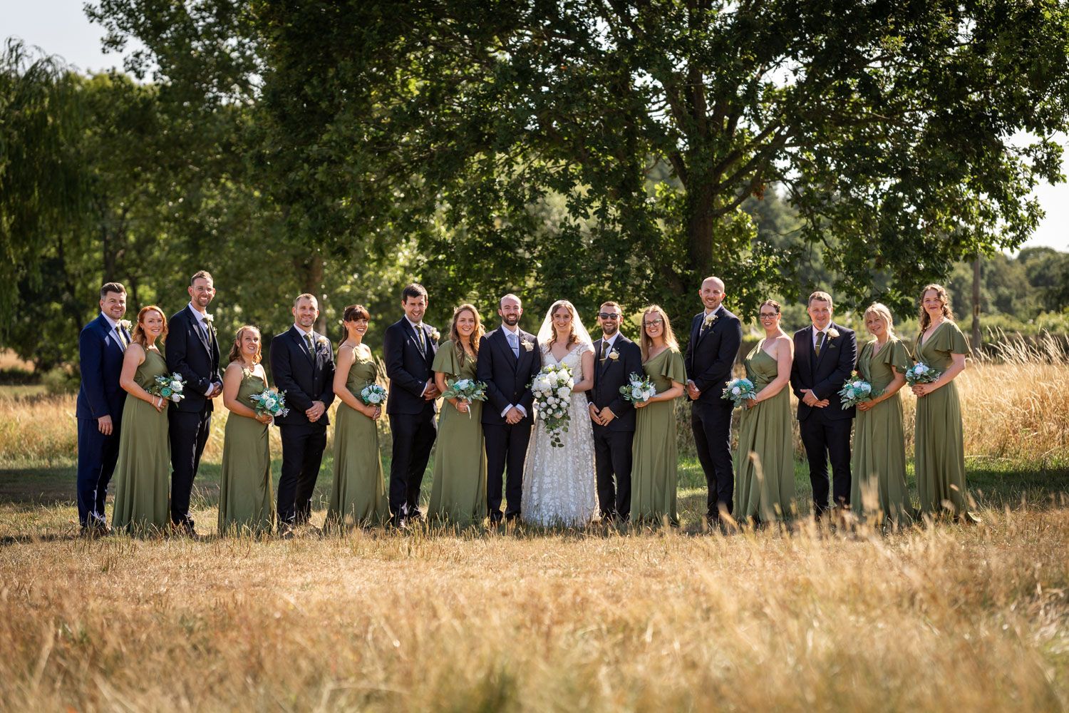 Wedding party posing in a golden field, bridesmaids in sage green dresses flanking the bride and groom under large trees.