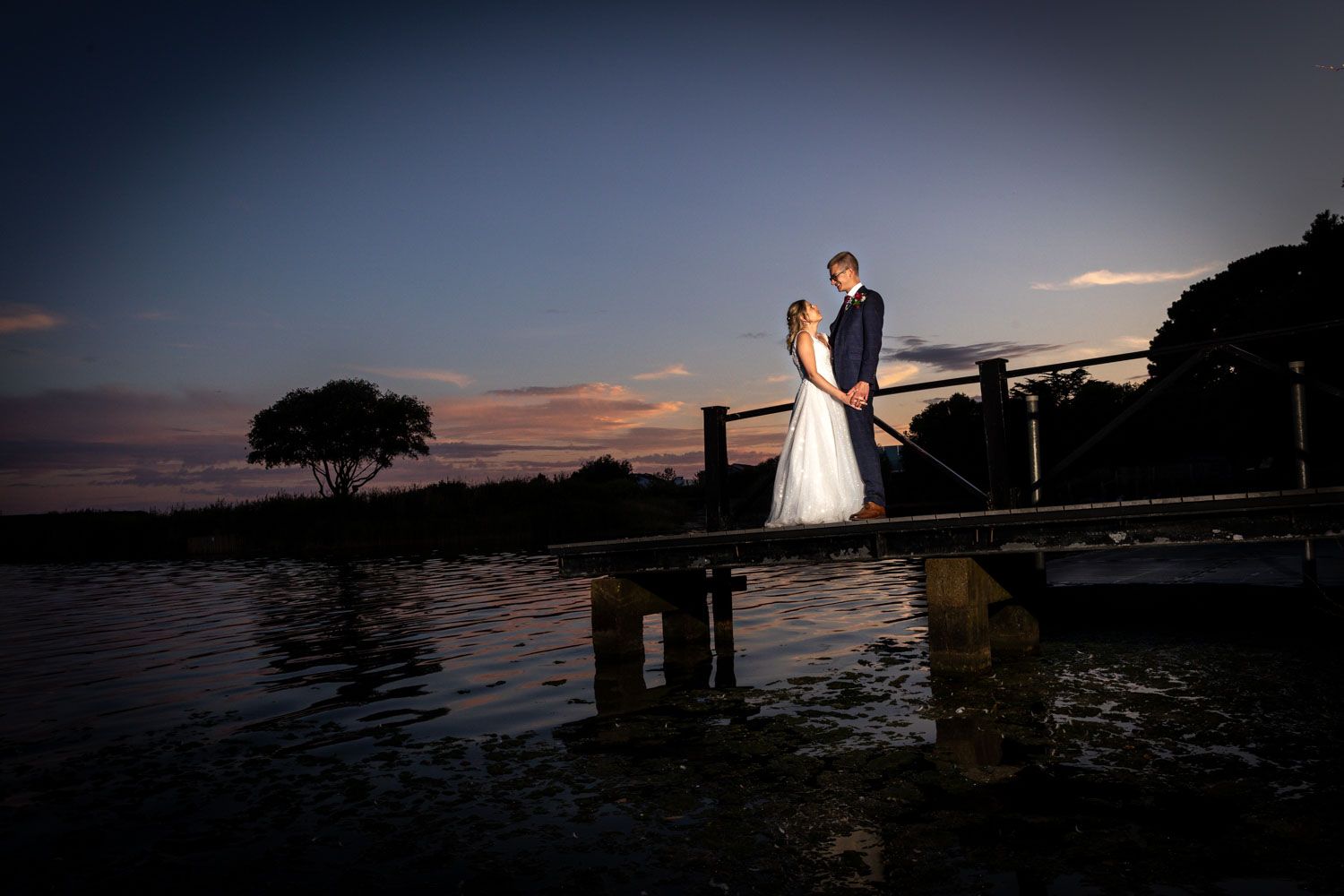 Bride and groom share a romantic moment on a dock at sunset with water reflections below.