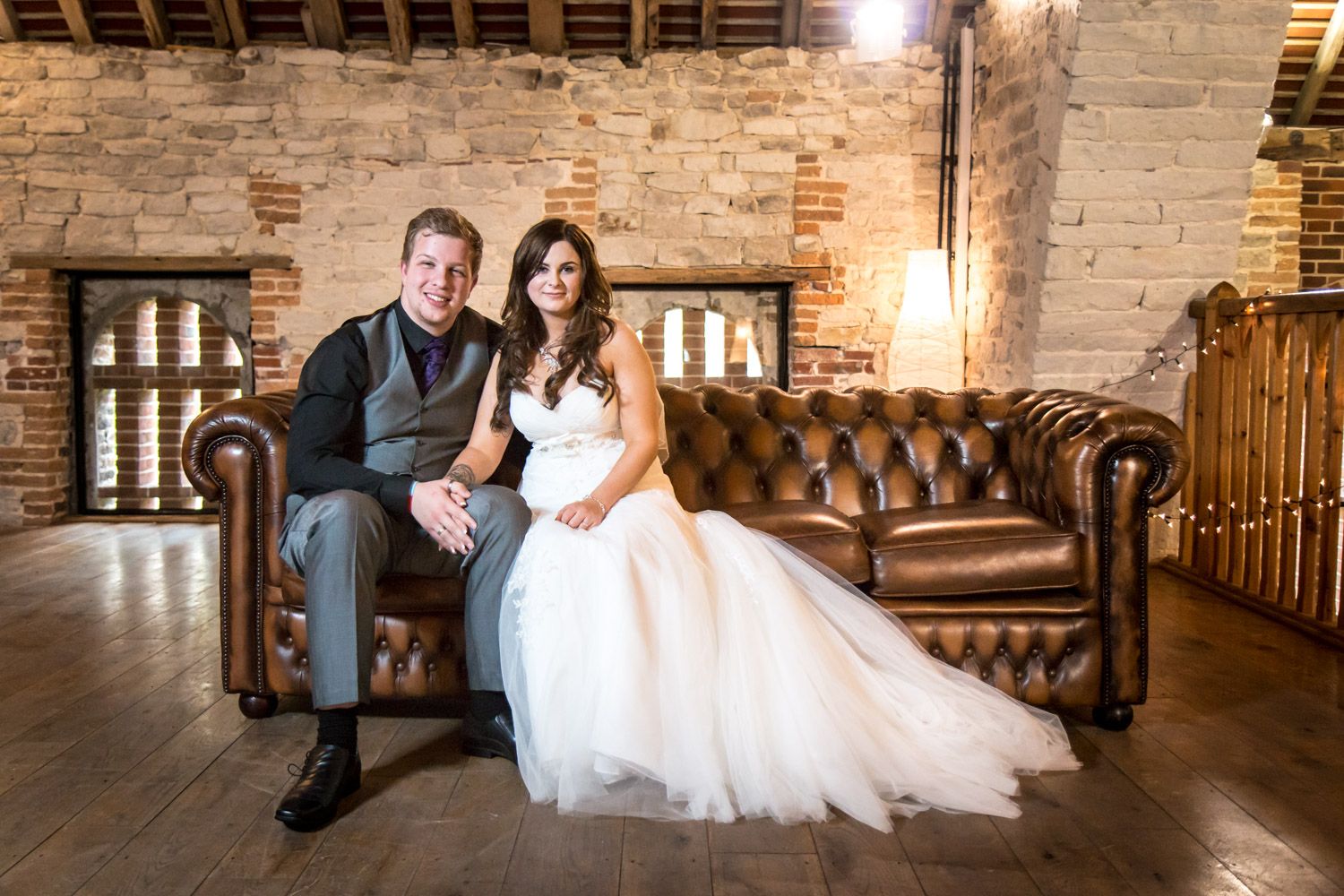 A couple sits together on a brown leather Chesterfield couch in a rustic brick-walled room with warm lighting.