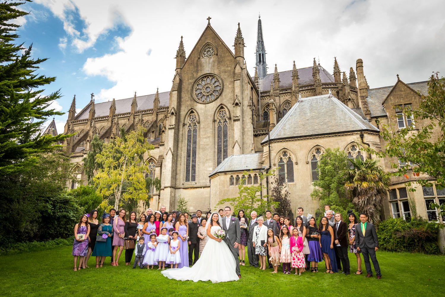 Wedding party poses in front of a grand Gothic cathedral surrounded by lush gardens.