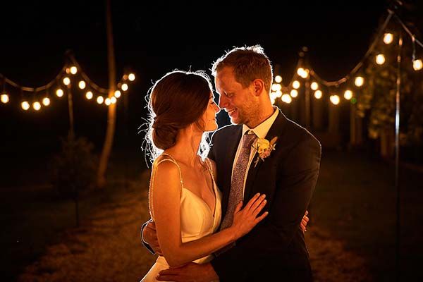 Bride and groom share a romantic moment under string lights at night during their wedding reception.