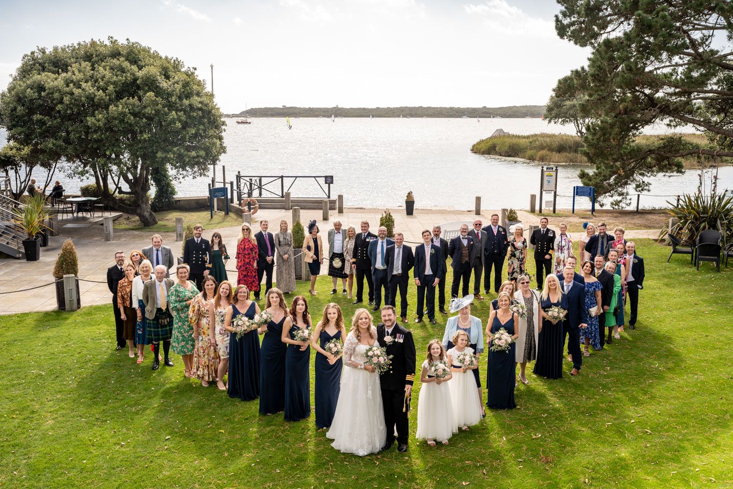 Wedding guests gathered in a heart shape formation on green lawn beside waterfront with boats and trees.
