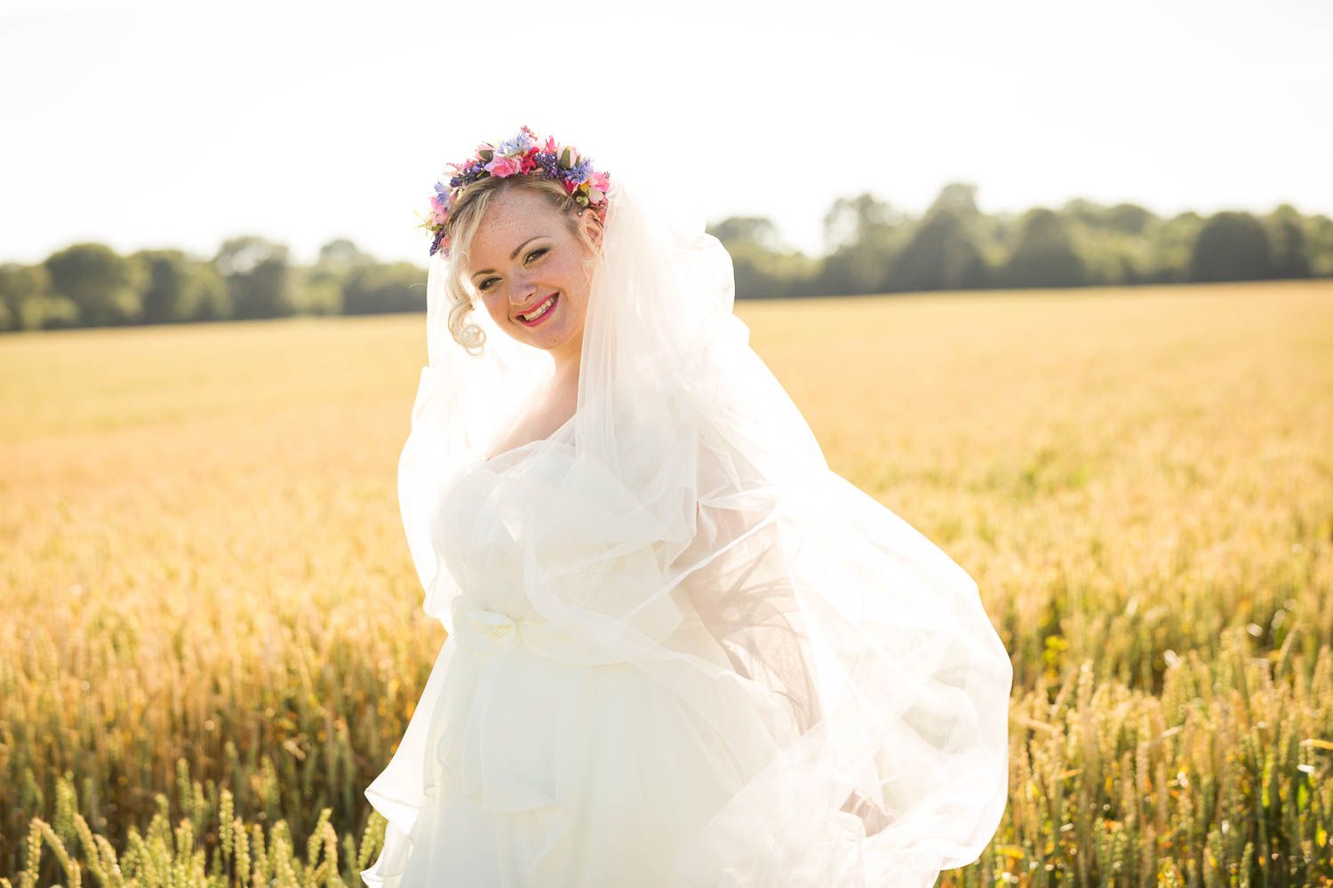 A bride in a flowing white wedding dress poses joyfully in a golden wheat field at sunset.
