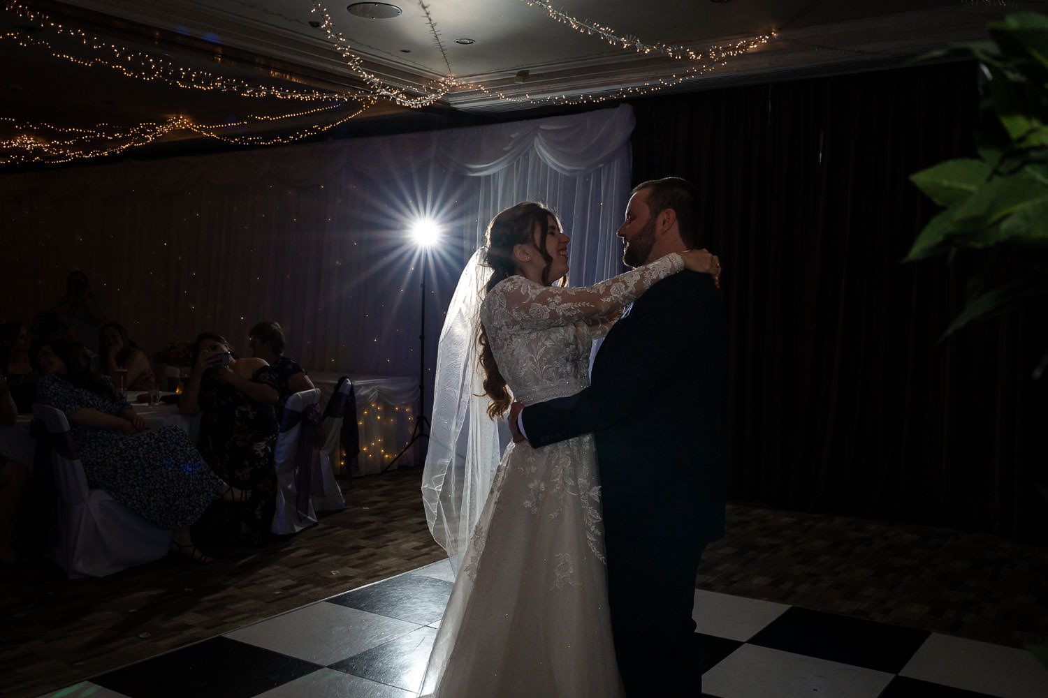 Newlywed couple shares their first dance under a spotlight with string lights glowing in the dark reception venue.