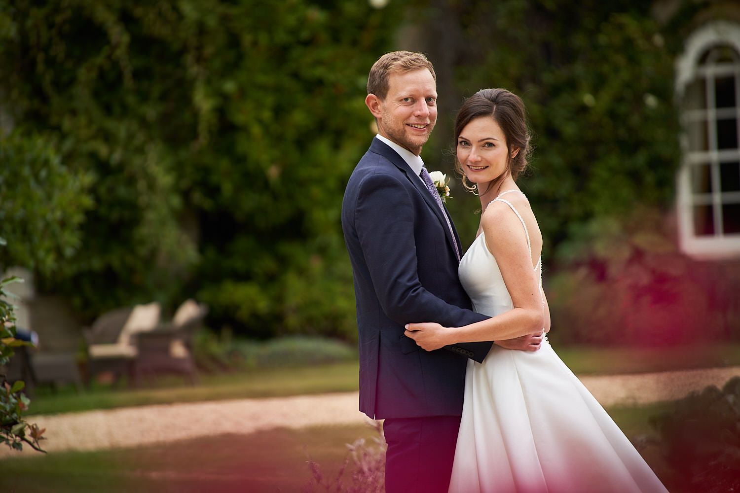 A couple in wedding attire pose together outdoors at a venue with scenic greenery and architecture in the background.