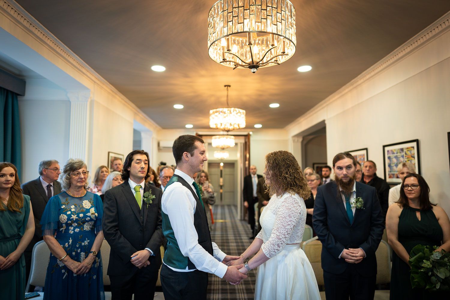 Wedding ceremony takes place in an elegant hallway with crystal chandelier and gathered guests.