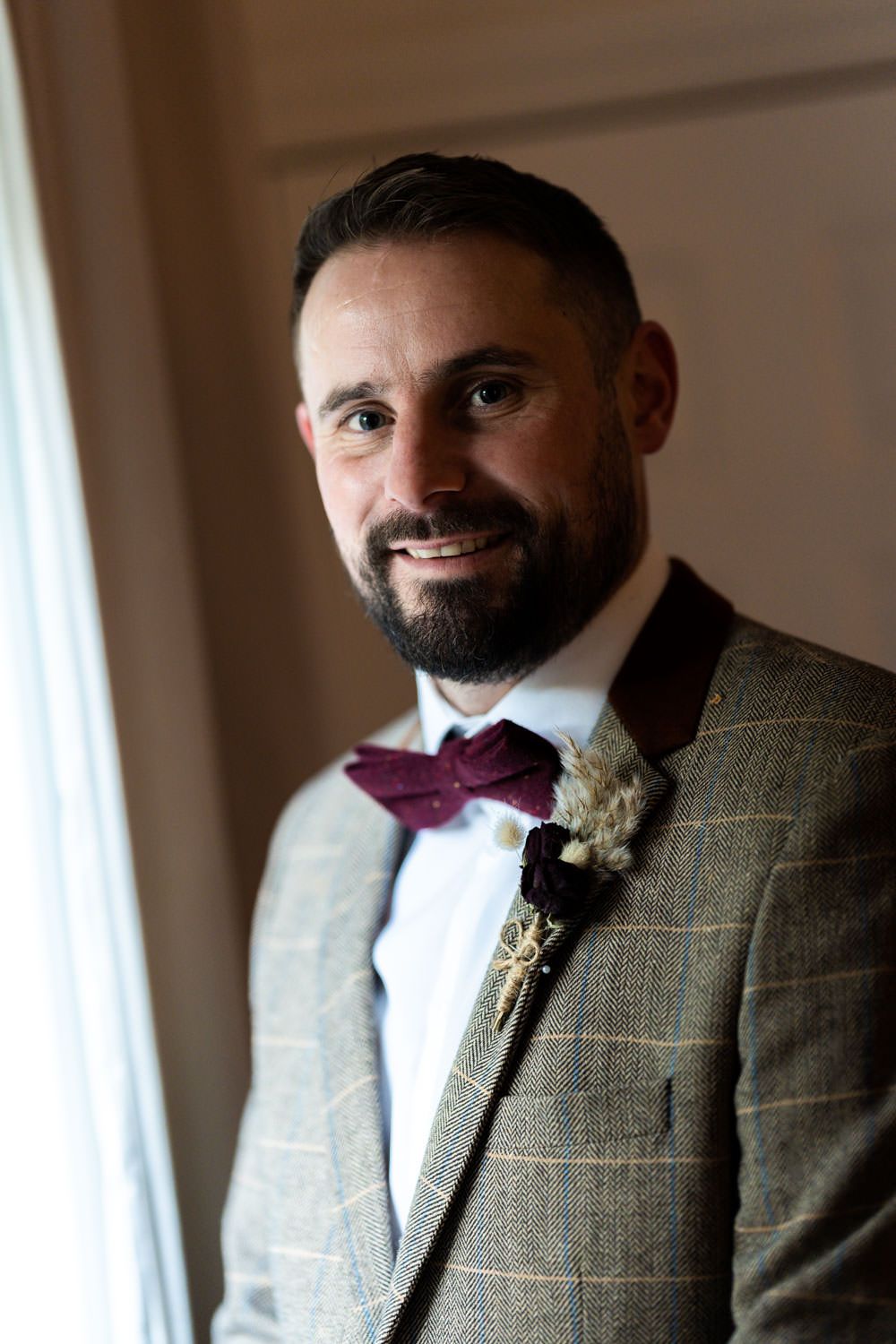 A groom in a checkered tweed suit and burgundy bow tie smiles while standing by a window.