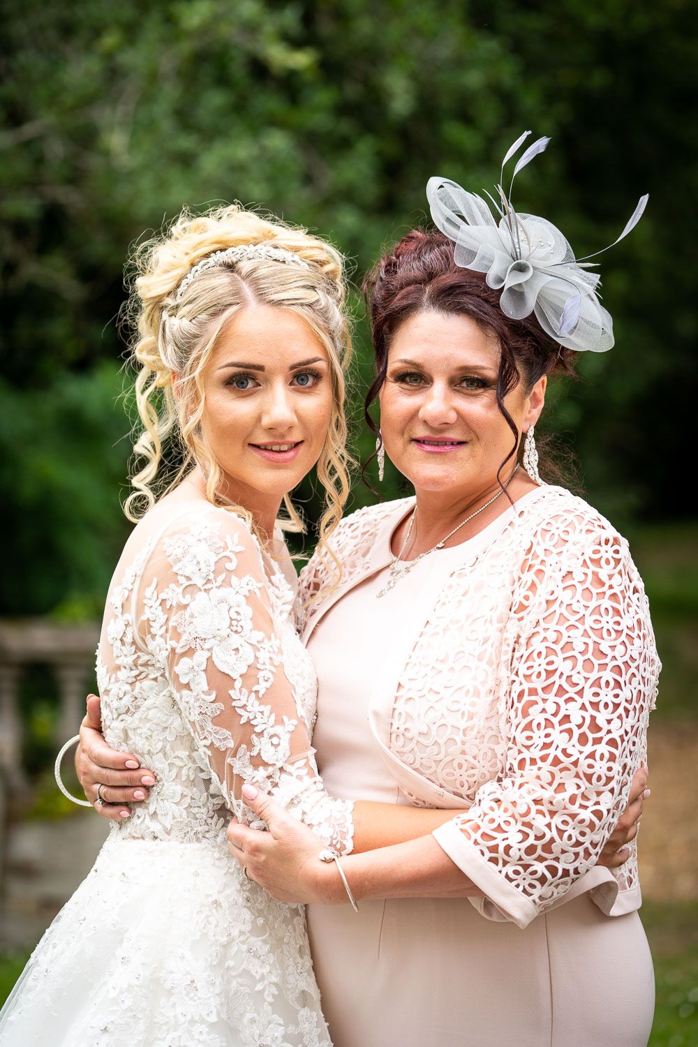 Two people in formal attire at a wedding wearing lace dresses, with one in white and one in blush pink with a fascinator.