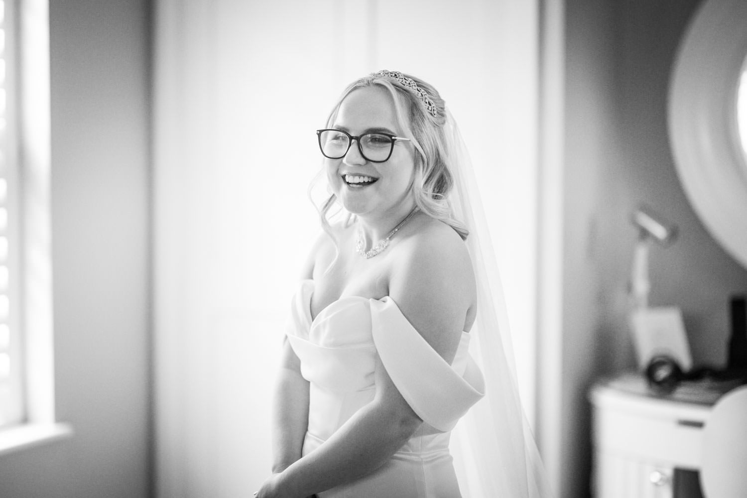 Black and white portrait of a bride in glasses wearing an off-shoulder wedding dress while looking back with a smile.