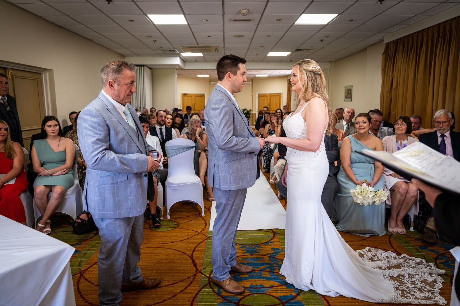 A wedding ceremony taking place in a bright hotel conference room with guests seated and watching the proceedings.