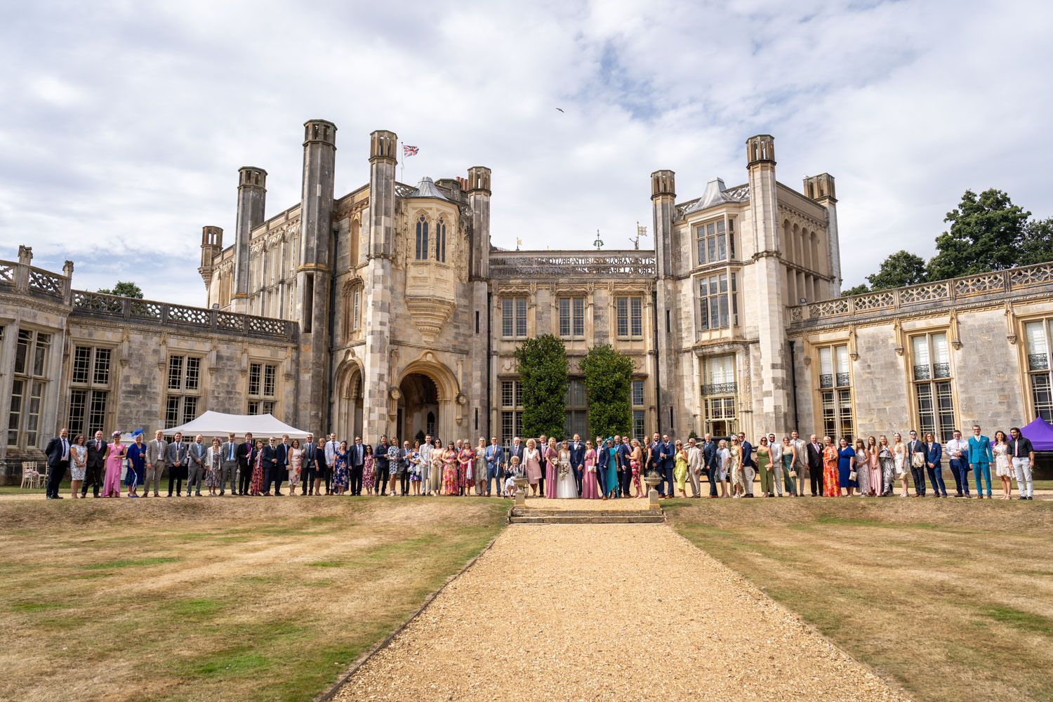 Wedding guests gather outside a grand historic manor house on a sunny day.