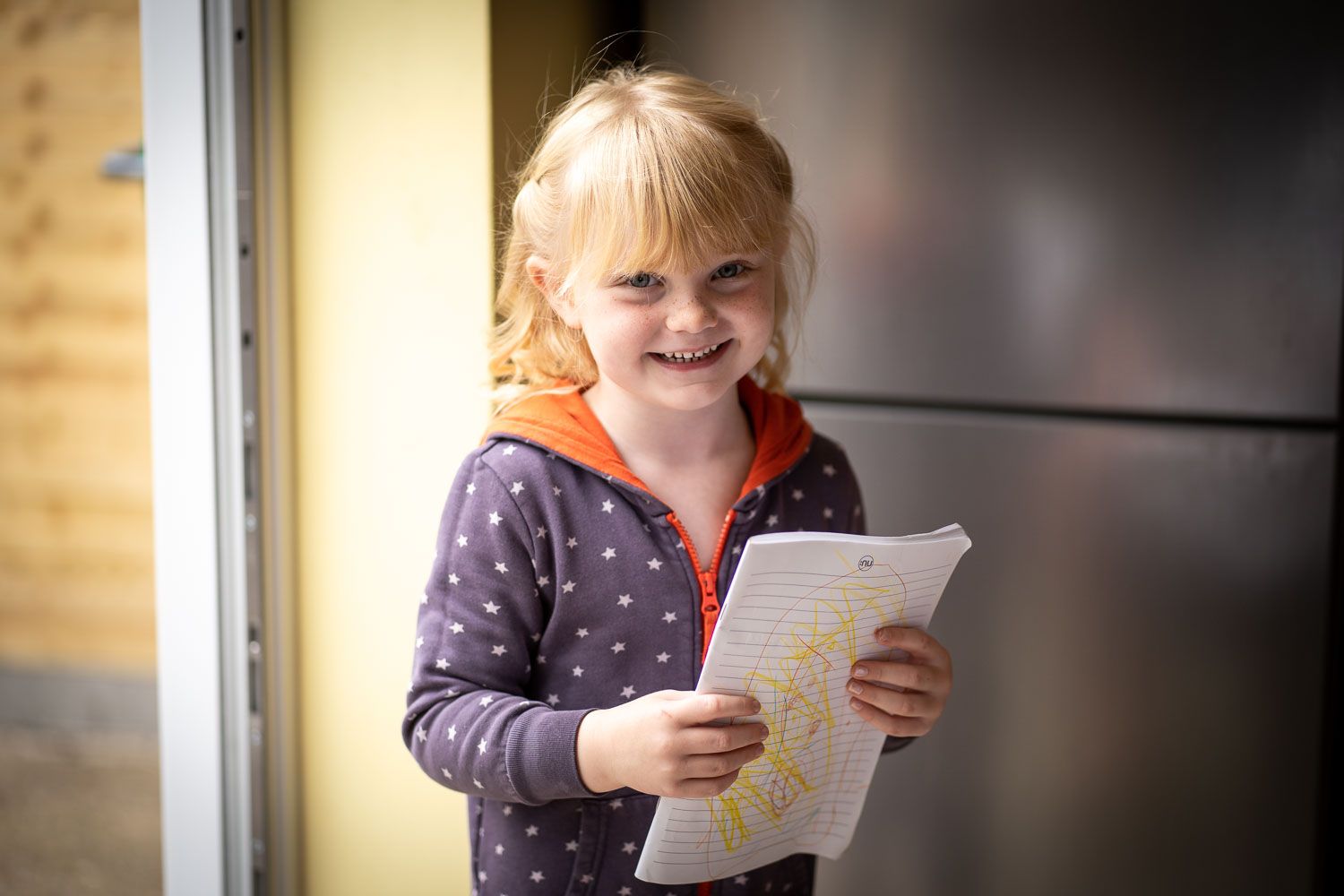 Young student in a polka dot sweater smiles while holding a sheet of paper in a sunny room.