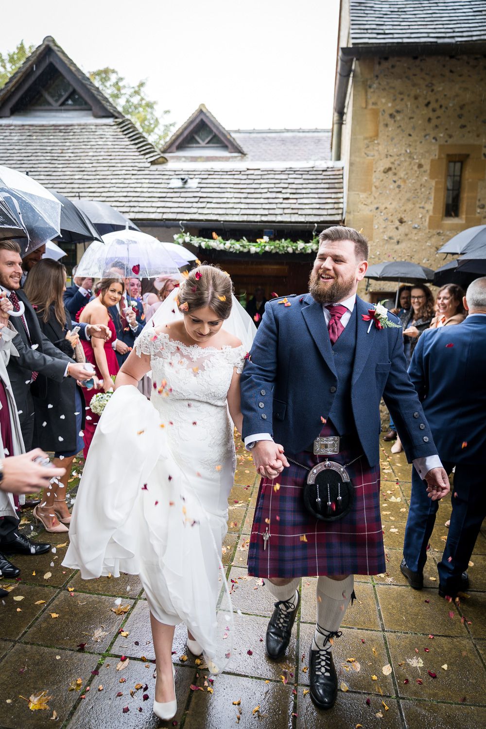 Newlyweds walk through confetti in the rain, with the groom wearing a traditional Scottish kilt and navy jacket.