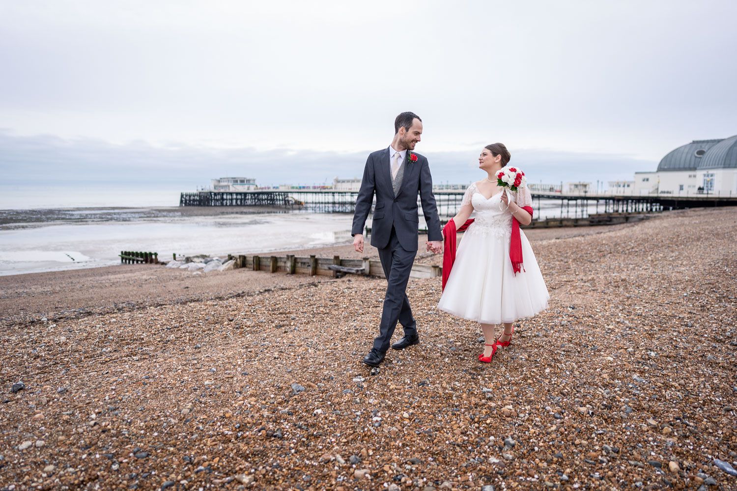 A couple strolls on a pebble beach with vintage red and white attire near a seaside pier on a cloudy day.