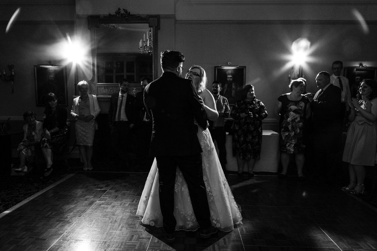 A couple shares their first dance at a wedding reception with guests watching from the dark dance floor.