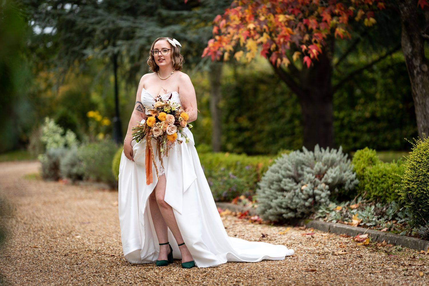 A joyful bride in a flowing white gown holds a vibrant fall bouquet on a garden path under autumn foliage.