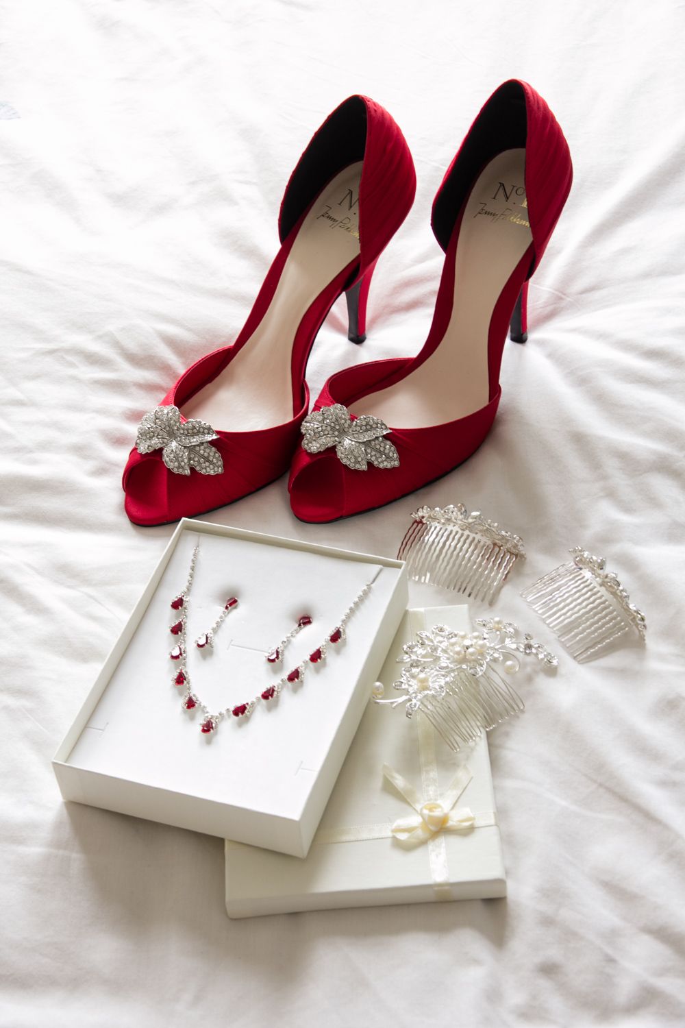 Elegant red satin heels with crystal butterfly embellishments displayed alongside delicate jewelry and accessories on white fabric.