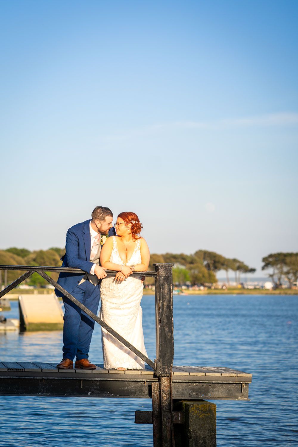 A romantic couple shares a tender moment on a wooden dock overlooking a serene waterfront at sunset.
