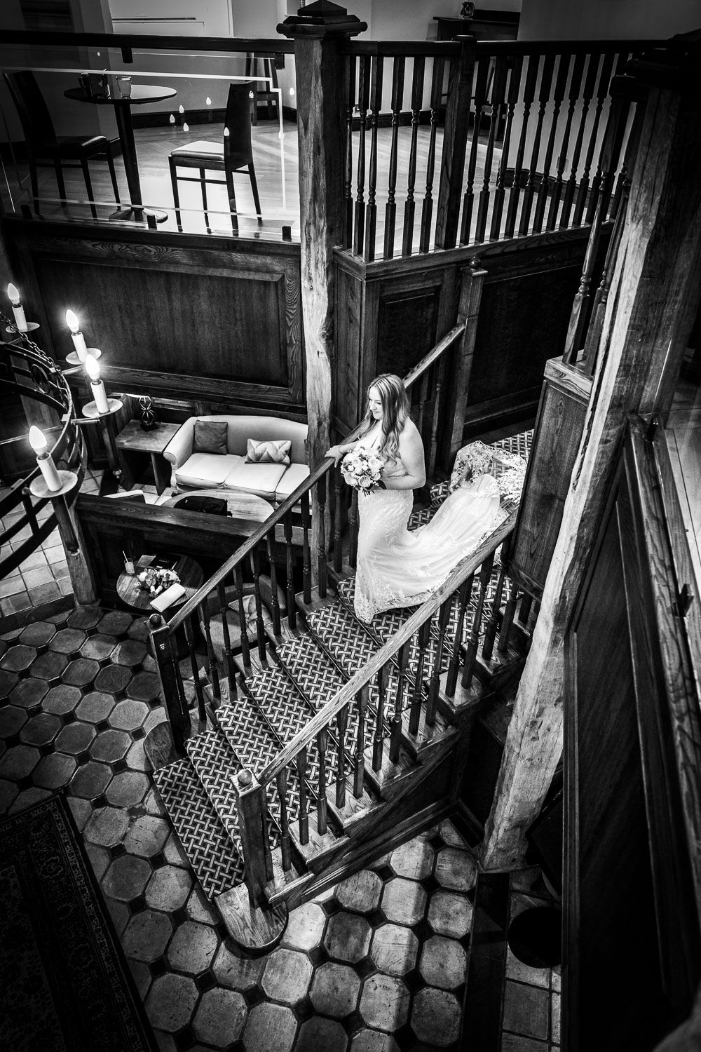 A dramatic black and white view of a vintage wooden staircase with Victorian details and patterned tile floor.