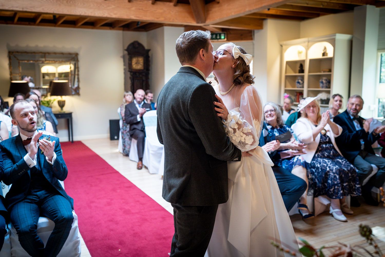 A couple shares their first kiss during a wedding ceremony in front of guests in an indoor venue with red carpet.