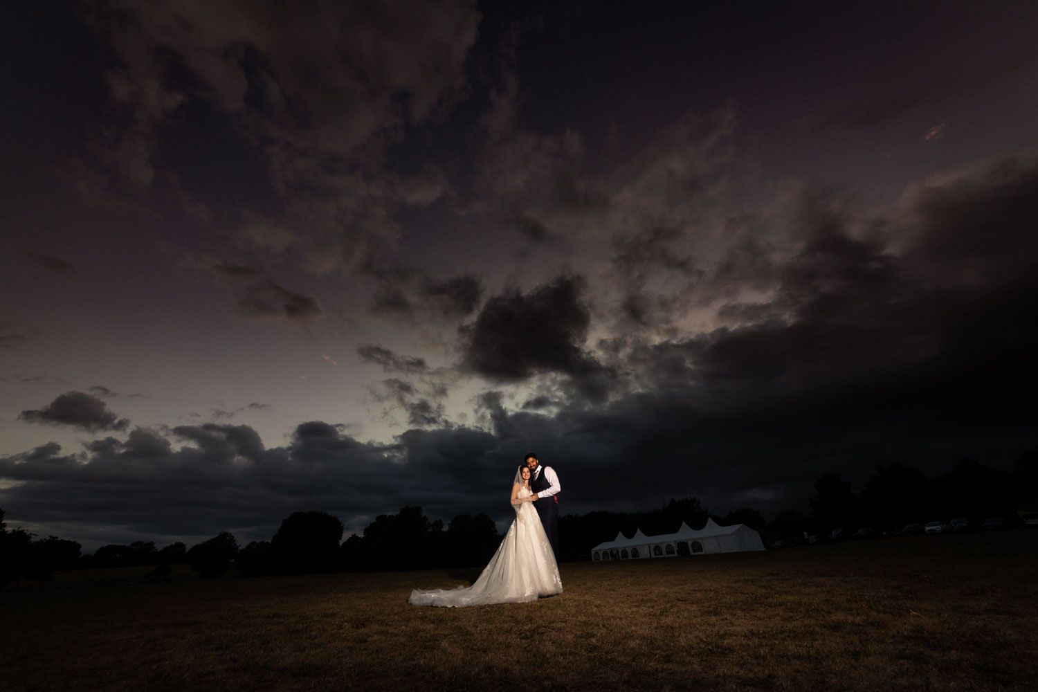 Silhouetted couple embracing in an open field under dramatic stormy sunset sky with dark clouds.