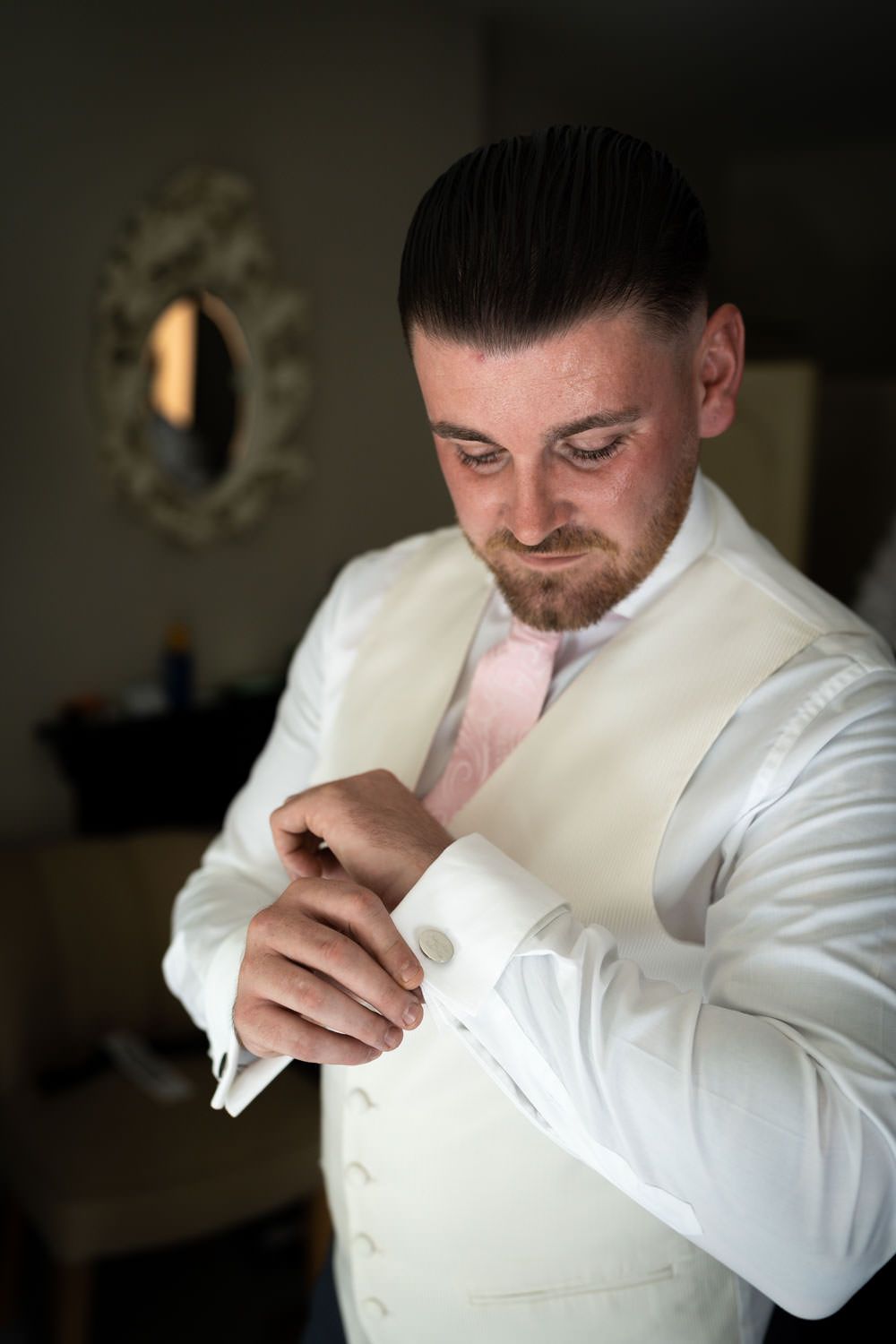 A groom in a white vest and pink tie adjusts his cuff while getting ready in a dimly lit room with an ornate mirror.