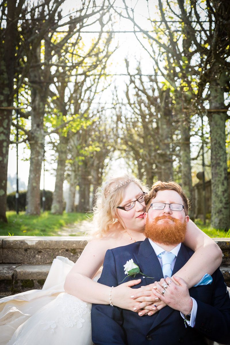 A couple embraces while sitting outdoors among a row of trees in a romantic wedding photo.