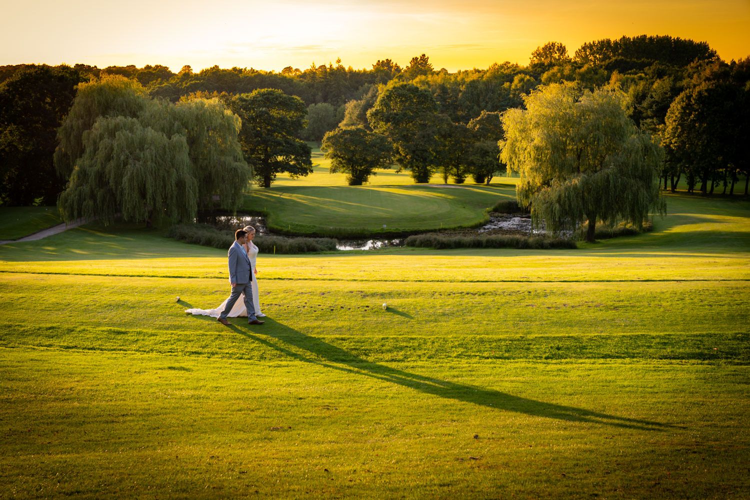 A couple embraces on a scenic golf course at sunset with weeping willow trees and golden light.
