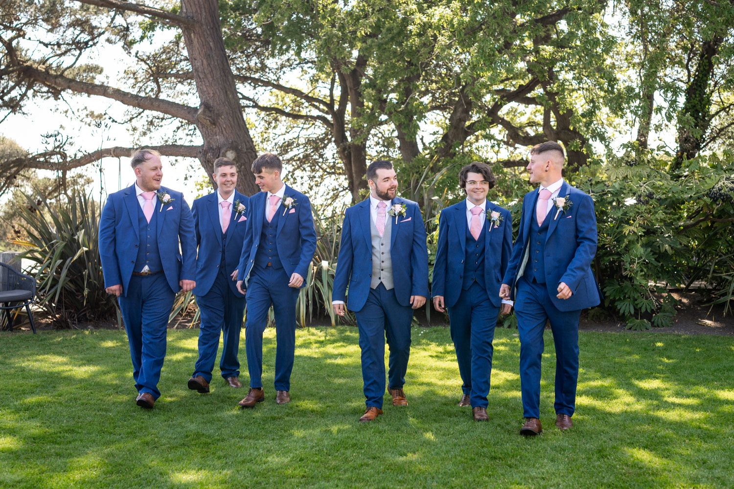 Groomsmen in matching royal blue suits stand together on green lawn during outdoor wedding ceremony.