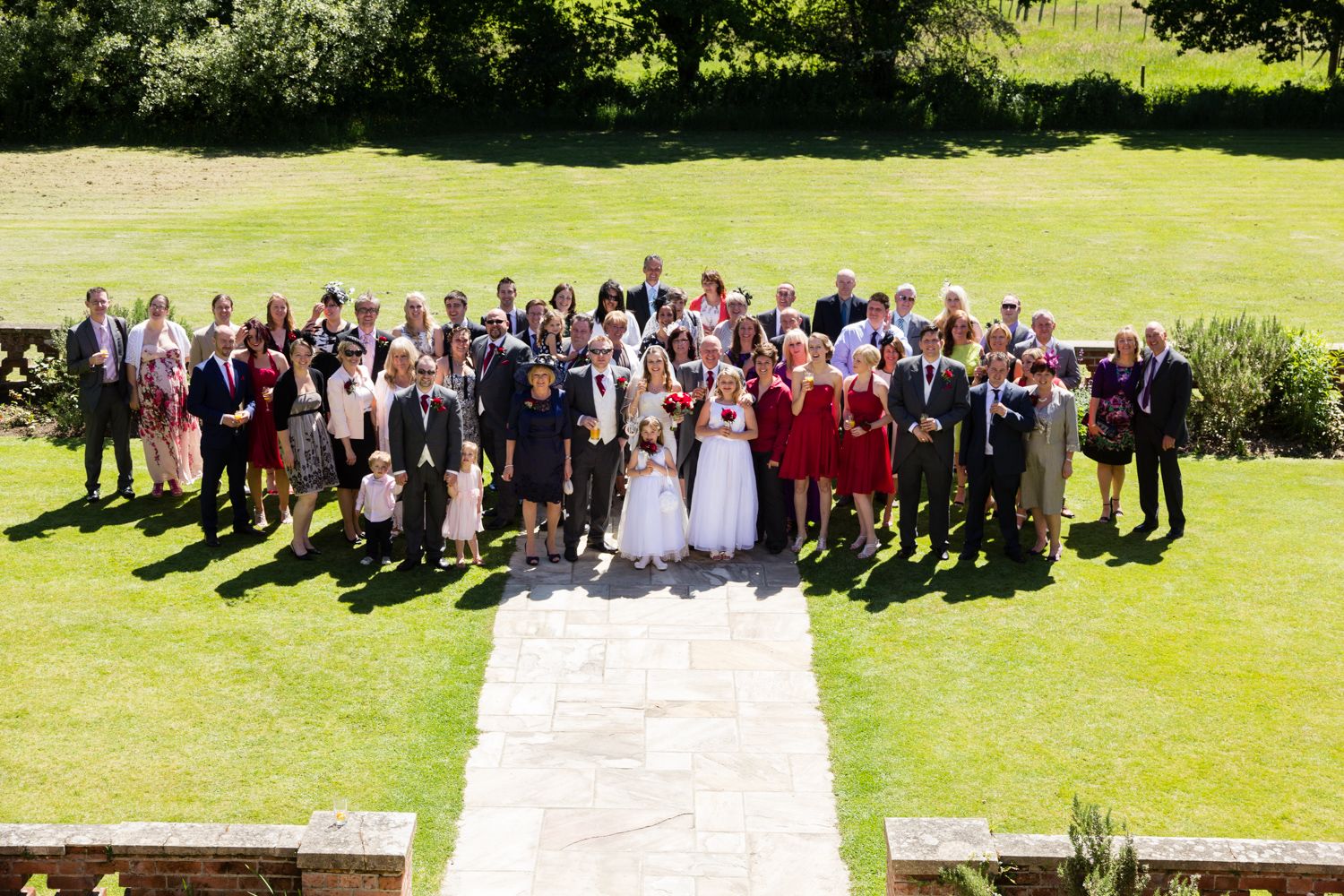 Large wedding party group photo taken outdoors on a sunny day with green lawn and gardens in the background.