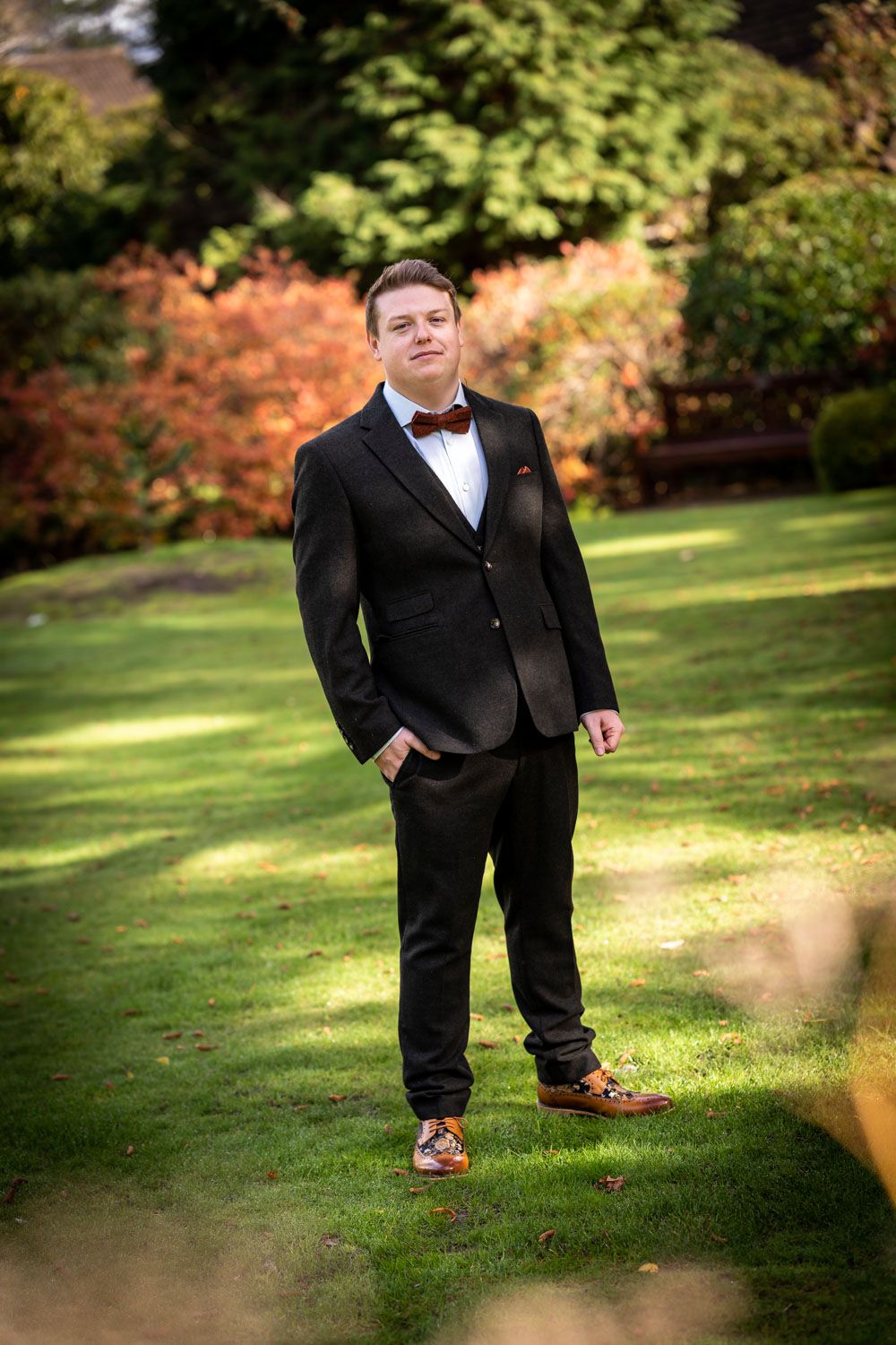 A person wearing a formal black tuxedo with bow tie stands on a lawn with autumn foliage in the background.