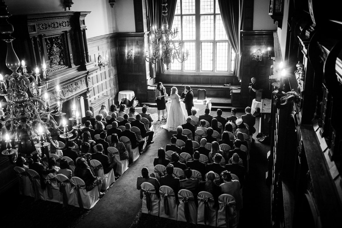 High angle black and white view of a wedding ceremony in a grand hall with chandeliers and rows of seated guests.