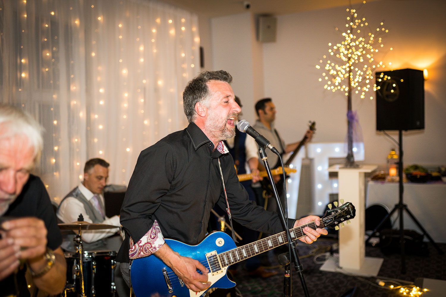 Live band performs at an indoor venue with a guitarist in black shirt playing electric guitar and string lights in background.
