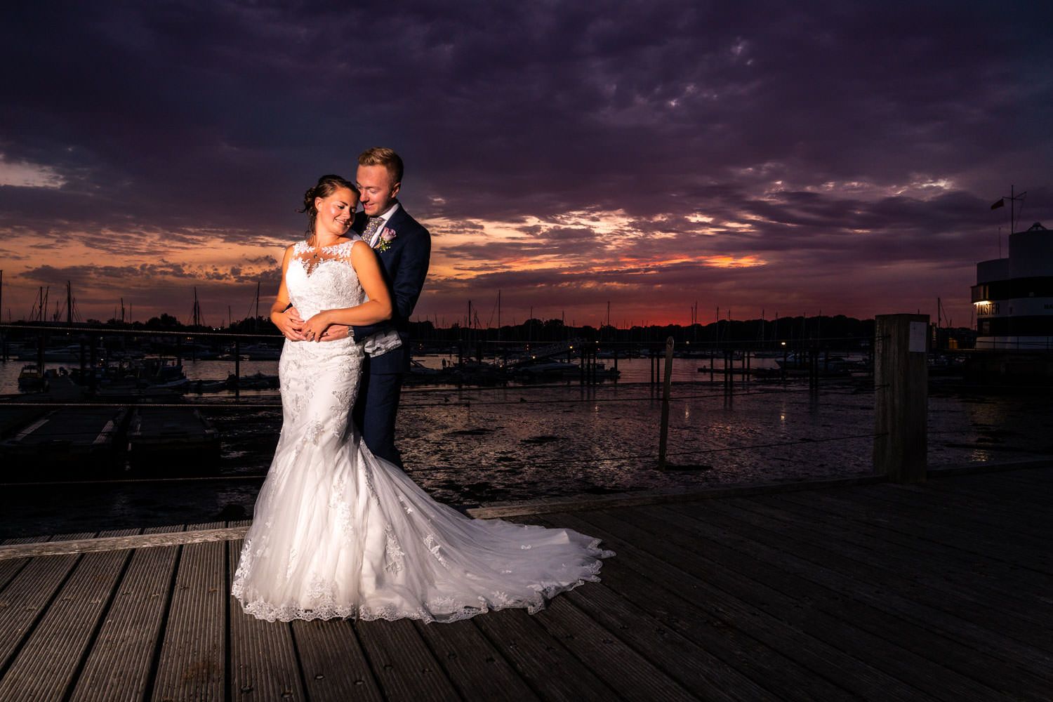 Bride in mermaid gown and groom embracing on dock with vibrant purple sunset over marina.