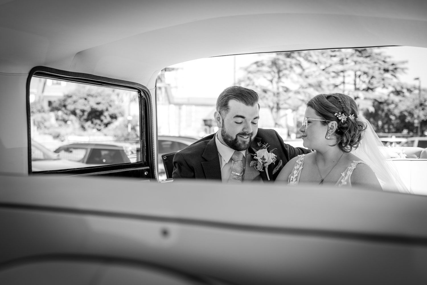 A vintage black and white photo shows a couple sitting together in the back seat of a classic car.