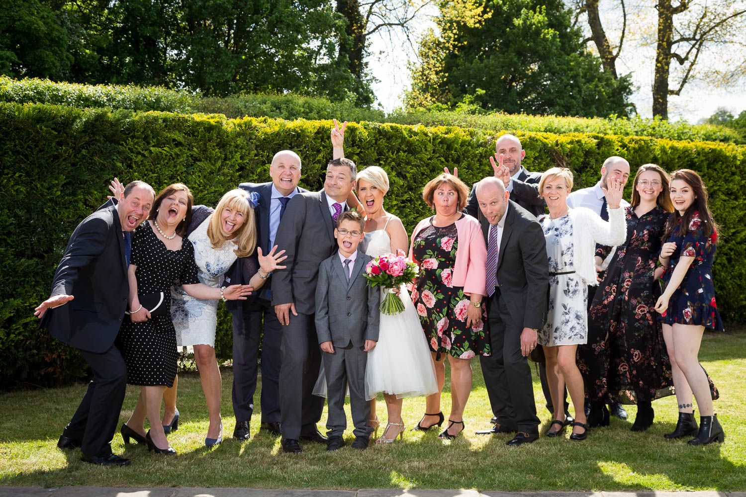 A group of wedding guests pose together outdoors in formal attire against a backdrop of green hedges on a sunny day.