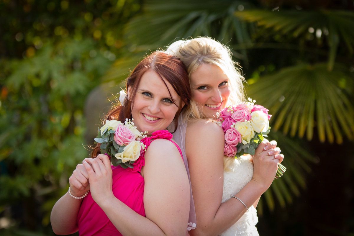 Two women in bright dresses pose back to back while holding pink and white flower bouquets in an outdoor garden setting.