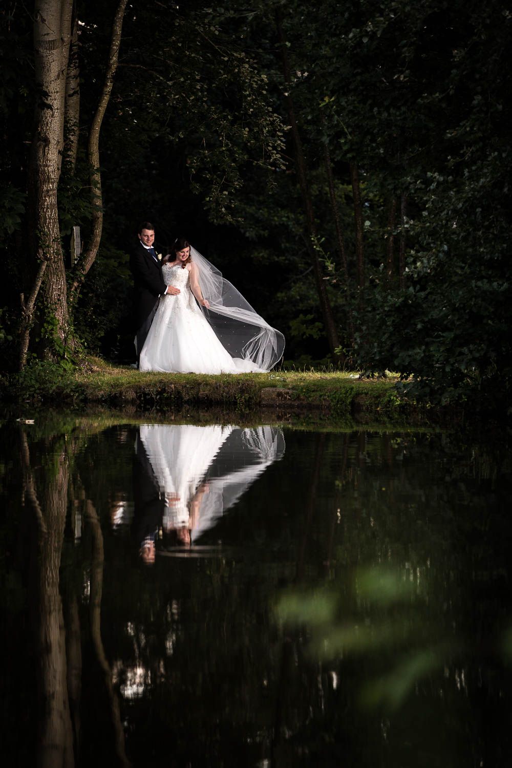 Romantic wedding couple stands by tranquil pond in dark forest, creating perfect mirror reflection in still water.