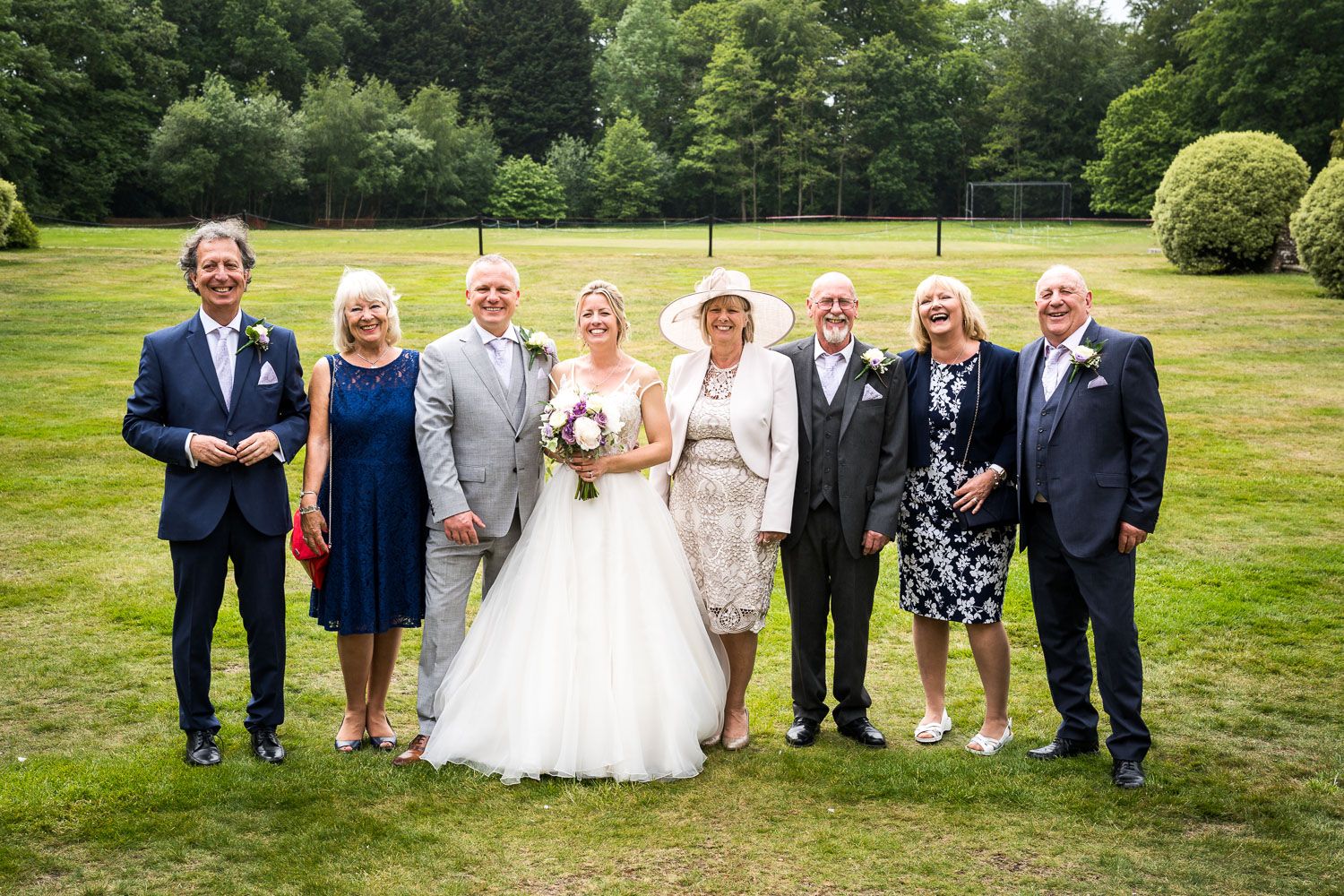 A group of formally dressed guests pose together on a lush green lawn during an outdoor summer wedding celebration.