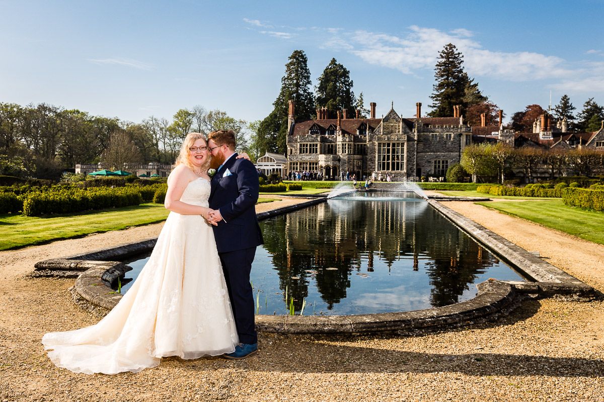 A couple celebrates their wedding at a grand estate with a reflecting pool and manicured gardens on a sunny day.