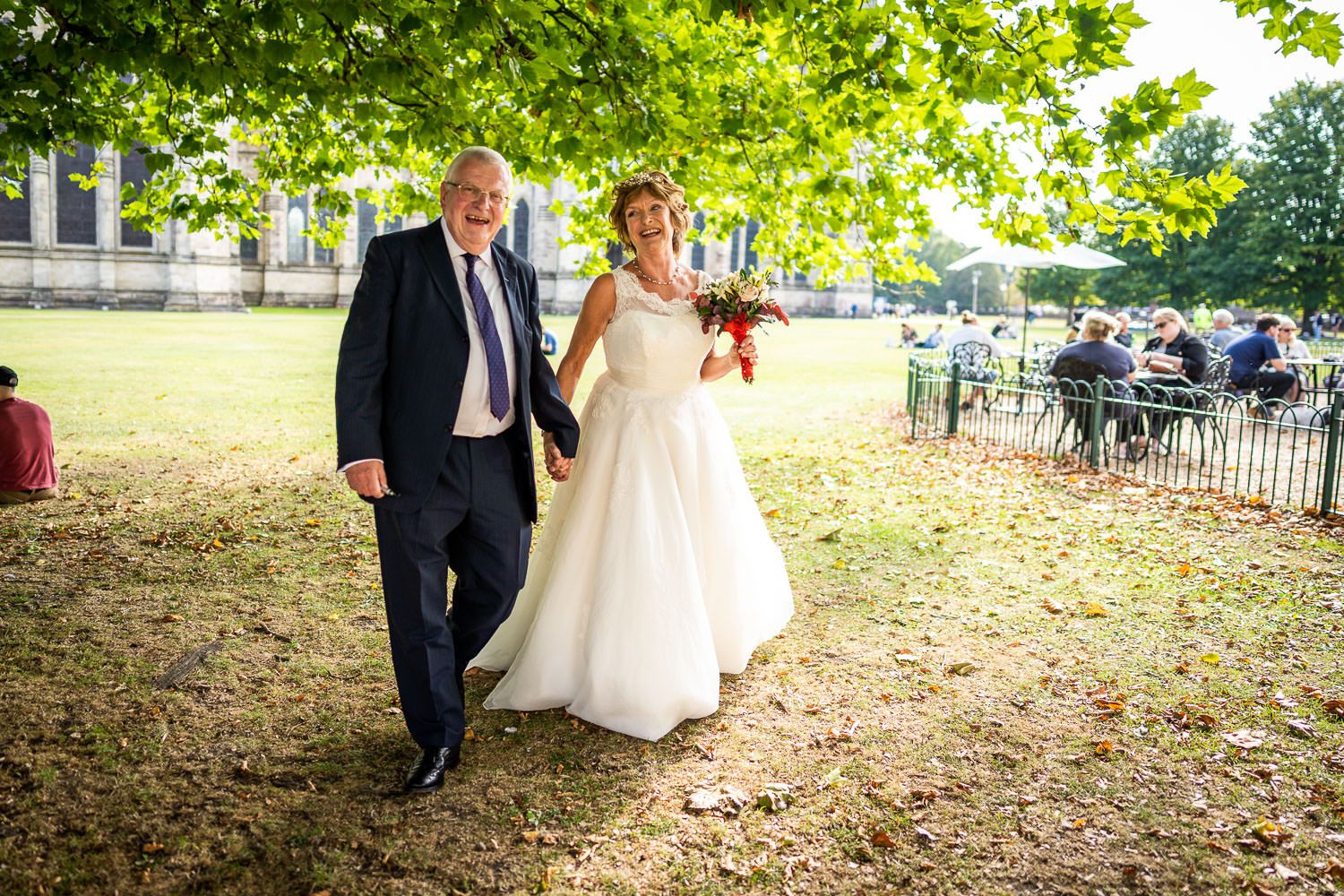 A couple walks together on grass beneath green trees on a sunny wedding day, captured in a candid moment.