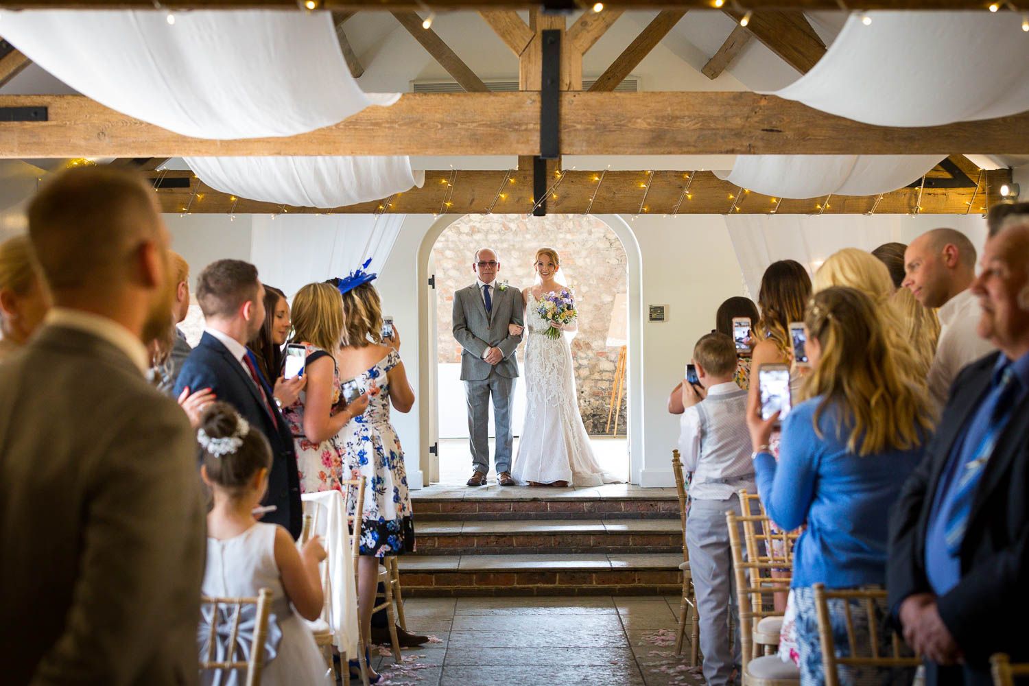 Indoor rustic barn wedding ceremony with exposed wooden beams and white fabric draping overhead during the vows.