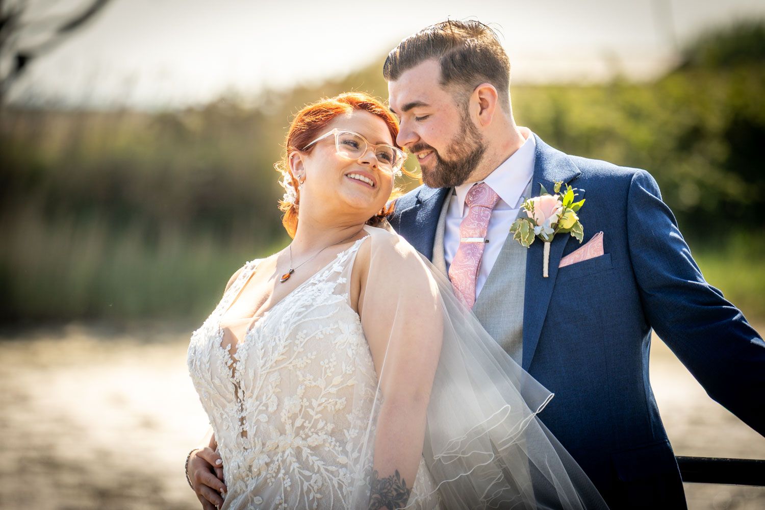 A couple in wedding attire embrace outdoors against a sunlit natural backdrop with a boutonniere on the navy suit.