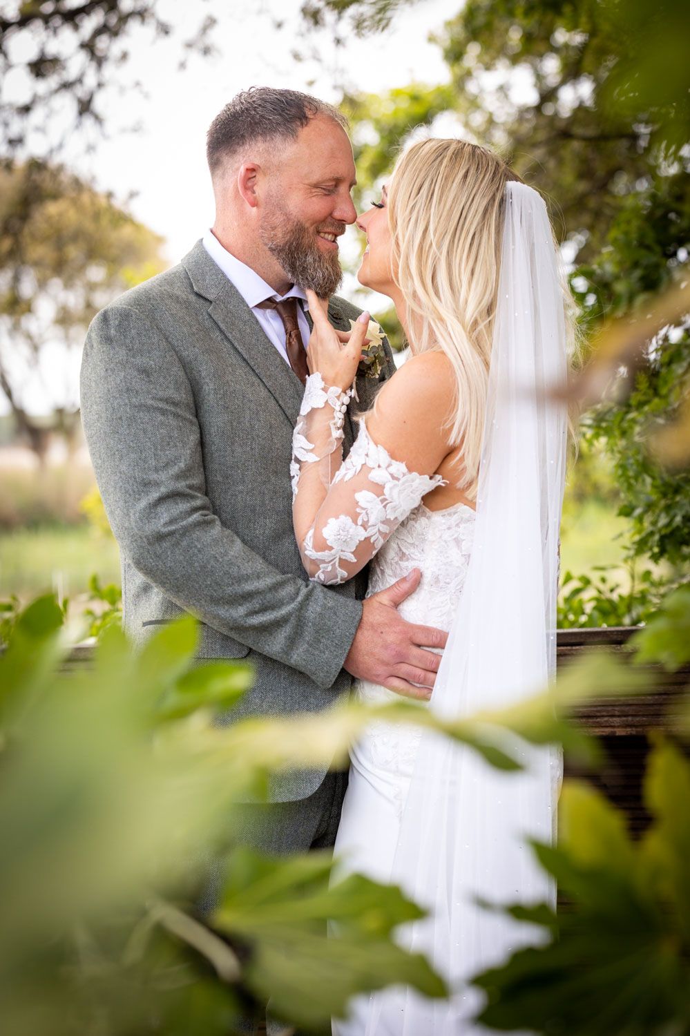 Bride and groom share a romantic moment surrounded by lush greenery, bride in lace gown with veil, groom in gray suit.