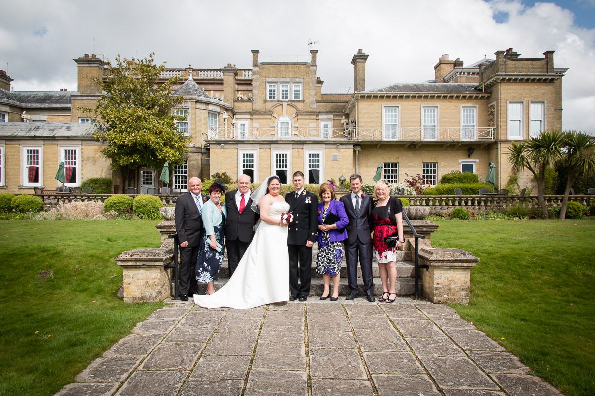 Wedding party gathered on stone steps in front of elegant English manor house with manicured lawn on sunny day.