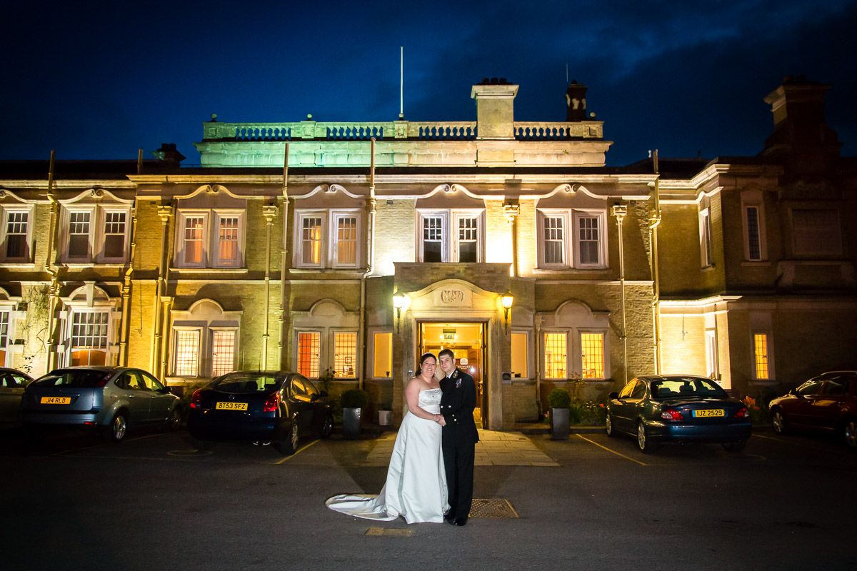 An elegantly lit Victorian mansion serves as a romantic wedding venue at night with a couple standing in front.