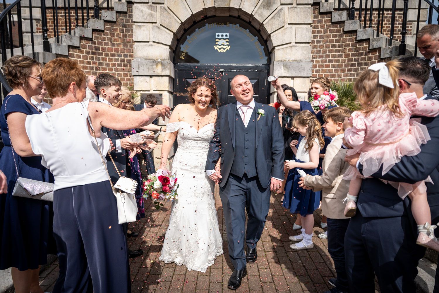 Wedding couple exits historic brick building while guests shower them with confetti and rose petals in celebration.